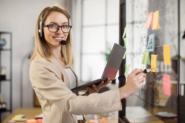 Wanita tersenyum dalam tulisan headset di papan kantor kaca — Stok Foto