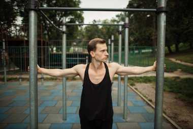 Kendine güveni tam genç kaslı bir adam dışarıda spor salonunda dikilirken kollarını açıyor..