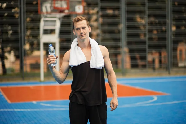 Homem bonito positivo em roupas esportivas posando na quadra de basquete ao ar livre com garrafa de água — Fotografia de Stock