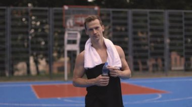 Omzunda havluyla atletik bir genç adam su içiyor ve bir sokak spor salonunda ya da açık hava basketbol sahasında yoğun bir idmandan sonra dinleniyor..