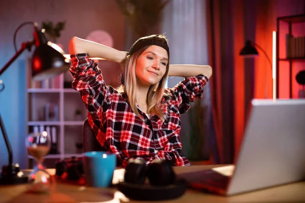 Attractive woman sitting at desk with hands behind head and looking at laptop screen. Caucasian lady in casual wear taking break during process hours at home.