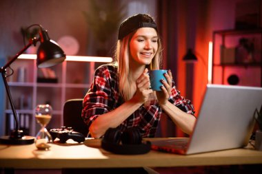 Likable young blond Caucasian woman sitting at desk with laptop and hot drink in her hands. Pleasant female in checkered shir using portable computer for work at dark room at home drinking coffee. clipart