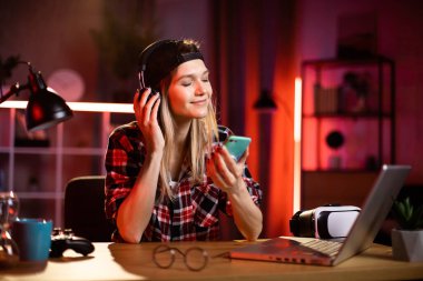 Sevimli bir kadın en sevdiği şarkıyı dinlemek için akıllı telefon ve kulaklık kullanıyor. Evde dizüstü bilgisayarla uğraşırken şapkalı genç bayan mola veriyor. Serbest ve teknoloji kavramı.