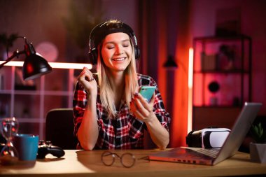 Sevimli bir kadın en sevdiği şarkıyı dinlemek için akıllı telefon ve kulaklık kullanıyor. Evde dizüstü bilgisayarla uğraşırken şapkalı genç bayan mola veriyor. Serbest ve teknoloji kavramı.