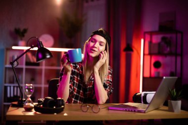 Kareli gömlekli, siyah şapkalı, telefonda konuşan, modern dekore edilmiş oturma odasındaki masada oturan oldukça memnun bir genç beyaz kadın. Çevrimiçi iş kavramı