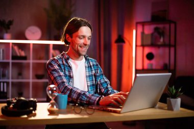 Evde ya da ofiste oturan ve kablosuz bilgisayarda yazan kareli gömlekli yakışıklı bir genç. Çekici erkek, akşam saatlerinde taşınabilir bilgisayarda çalışıyor..