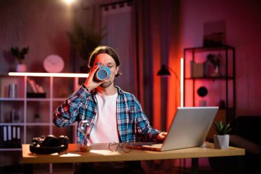 Akşamları masa başında oturan beyaz bir adam video aracılığıyla iş arkadaşlarıyla konuşuyor. Gündelik giysiler içinde genç bir erkek, modern dizüstü bilgisayarını evde uzaktan çalışmak için kullanıyor..