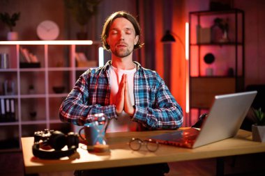 Kareli gömlekli gülümseyen beyaz adamın portresi. Akşam evde yoga egzersizi yapıyor. Uzaktan kumanda sırasında meditasyon için ara veren mutlu genç adam..