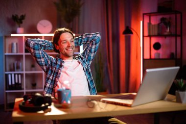 Kareli gömlekli hoş bir adamın portresi. Elleri arkada, Tabel 'de oturmuş, modern dizüstü bilgisayarla gülümsüyor ve kameraya bakıyor. Evdeki karanlık atmosfer. Akşam saatlerinde uzak çalışma.