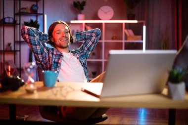 Kareli gömlekli hoş bir adamın portresi. Elleri arkada, Tabel 'de oturmuş, modern dizüstü bilgisayarla gülümsüyor ve kameraya bakıyor. Evdeki karanlık atmosfer. Akşam saatlerinde uzak çalışma.