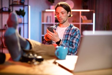 Kareli gömlekli yakışıklı genç adam ayaklarını modern akıllı telefonuyla masaya koydu. Akşam saatlerinde internette cep telefonuyla internette sörf yapan güzel bir adam..