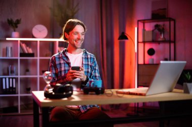 Akıllı telefon ve dizüstü bilgisayar kullanan beyaz bir adam. Kareli gömlekli. Evde dizüstü bilgisayarla uğraşırken ara veren genç adam. Serbest ve teknoloji kavramı.