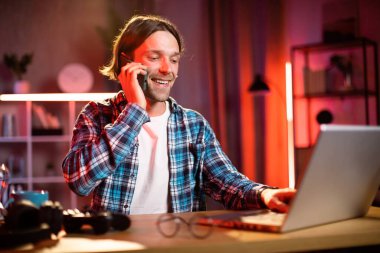 Sarışın ve kareli gömlekli çekici bir adam masada oturmuş dizüstü bilgisayarda yazı yazıyor ve telefonda konuşuyor. Odaklanmış mavi gömlekli erkek karanlık ofiste çalışmak için taşınabilir bilgisayar kullanıyor..