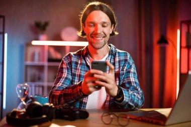 Akşamları evde akıllı telefon kullanan kareli gömlekli beyaz bir adamın portresi. Modern aletlerle yaşam tarzları.