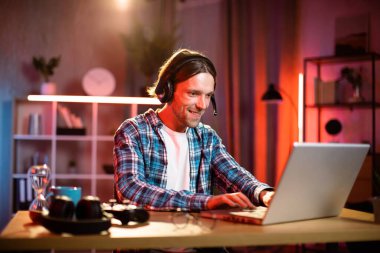 Kulaklıkla odaklanmış erkek yönetici. Bilgisayardaki video görüşmesiyle iş toplantısına öncülük ediyor. Genç adam evdeki masada otururken konuşuyor ve el kol hareketi yapıyor. Uzaktan çalışıyor.