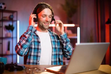 Kulaklık takmış genç bir beyaz adamın portresi masasında oturuyor ve evde uzaktan çalışmak için modern bir dizüstü bilgisayar kullanıyor. Erkek serbest çalışan ciddi bir yüz ifadesi olan dizüstü bilgisayara bakıyor..