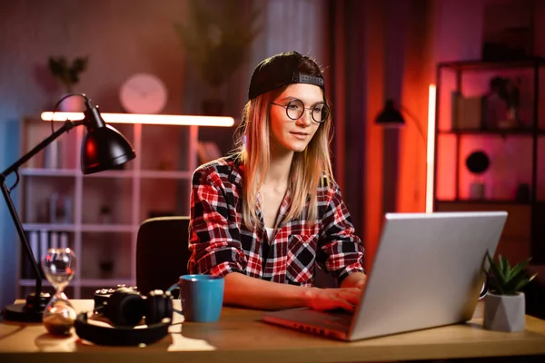 Kareli gömlekli ve gözlüklü genç bir kadın akşam vakti masada otururken modern dizüstü bilgisayarda çalışıyor. Uzaktan kumandalı iş. Serbest ve insan konsepti.