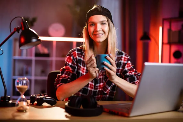 Smiling Caucasian woman drinking hot coffee while using laptop computer at dark office or home at night. Pretty female software developer using modern laptop for remote work at home.