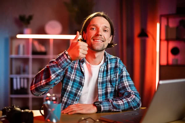 Çekici bir adam masada oturuyor, elleri göğsünde çaprazlanmış ve kameraya bakıyor. Gündelik giysiler içinde beyaz erkek. İşlem saatleri boyunca evde başparmağını gösteriyor..