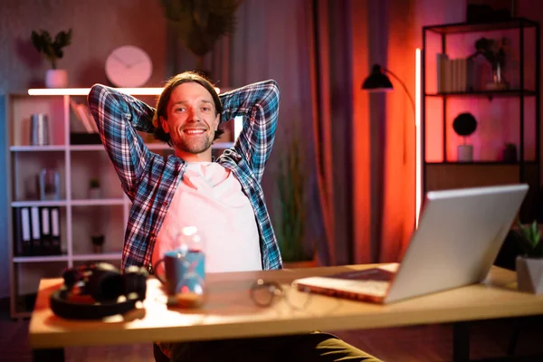 Kareli gömlekli hoş bir adamın portresi. Elleri arkada, Tabel 'de oturmuş, modern dizüstü bilgisayarla gülümsüyor ve kameraya bakıyor. Evdeki karanlık atmosfer. Akşam saatlerinde uzak çalışma.