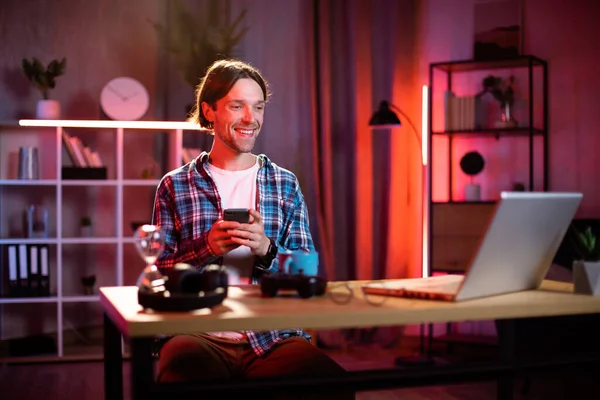 Akıllı telefon ve dizüstü bilgisayar kullanan beyaz bir adam. Kareli gömlekli. Evde dizüstü bilgisayarla uğraşırken ara veren genç adam. Serbest ve teknoloji kavramı.