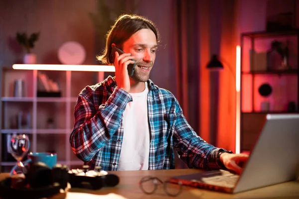 Sarışın ve kareli gömlekli çekici bir adam masada oturmuş dizüstü bilgisayarda yazı yazıyor ve telefonda konuşuyor. Odaklanmış mavi gömlekli erkek karanlık ofiste çalışmak için taşınabilir bilgisayar kullanıyor..