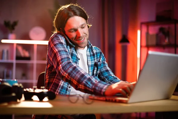 Sarışın ve kareli gömlekli çekici bir adam masada oturmuş dizüstü bilgisayarda yazı yazıyor ve telefonda konuşuyor. Odaklanmış mavi gömlekli erkek karanlık ofiste çalışmak için taşınabilir bilgisayar kullanıyor..