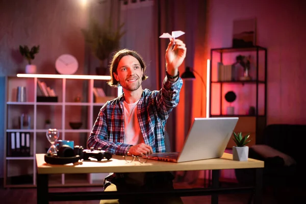 Gündelik kıyafetlerle gezen mutlu genç adam dizüstü bilgisayarda çalışırken kağıt uçak fırlatırken. Gülümseyen beyaz erkek akşam vakti masada oturuyor ve eğleniyor..