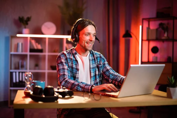 Kulaklıkla odaklanmış erkek yönetici. Bilgisayardaki video görüşmesiyle iş toplantısına öncülük ediyor. Genç adam evdeki masada otururken konuşuyor ve el kol hareketi yapıyor. Uzaktan çalışıyor.