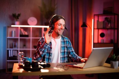 Kulaklık takmış genç beyaz bir adam dizüstü bilgisayarla masada oturuyor, video görüşmesi ya da çevrimiçi eğitim dersi alıyor. Çevrimiçi konuşma için modern cihazlar kullanan işadamı, merkez ofisten online çalışıyor.