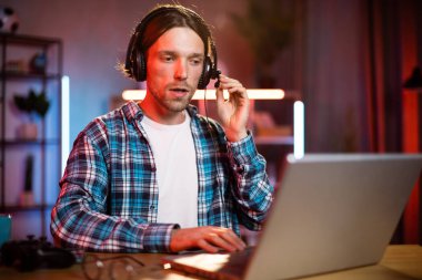 Kulaklık takmış genç bir beyaz adamın portresi masasında oturuyor ve evde uzaktan çalışmak için modern bir dizüstü bilgisayar kullanıyor. Erkek serbest çalışan ciddi bir yüz ifadesi olan dizüstü bilgisayara bakıyor..