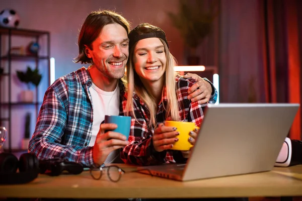 Young beautiful happy couple sitting at wooden table at night time in living room and watching movie and drink hot drinks tea or coffee in mugs using laptop. Concept of evening leisure and technology.