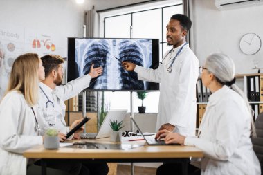 Tıbbi personel toplantısı sırasında röntgen cihazıyla dijital ekrana bakan dört çok kültürlü doktor. Afrikalı Amerikalı doktor, beyaz sakallı meslektaşıyla hasta teşhisi hakkında tartışıyor..