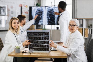 Yetenekli kadın terapistler gülümseyip ellerinde röntgen taramasıyla kameraya bakarken erkek iş arkadaşları büyük ekranın yanında çalışıyor. Bir grup çok ırklı doktor hasta sağlık sorunlarını tartışıyor.