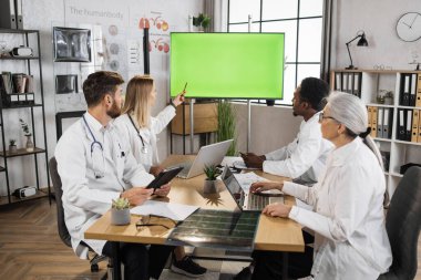 Çok kültürlü tıp uzmanlarından oluşan bir ekip modern aletlerle masada oturuyor ve krom ekranlı büyük ekrana bakıyor. Hastane ofisinde doktorların konferans toplantısı..