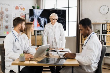 Deneyimli kıdemli kadın doktorlar, konferans sırasında röntgen taraması ve kalp atışı hızlarıyla monitörde yürüyorlar. Bir grup tıp çalışanı modern yönetim kurulunda beyin fırtınası yapıyor..