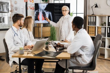 Kalifiye kadın doktor, kurul odasında üç çok ırklı stajyere seminer veriyor. Dijital ekranın yanında duran yaşlı kadın göğüs röntgeni sonuçlarını açıklıyor..