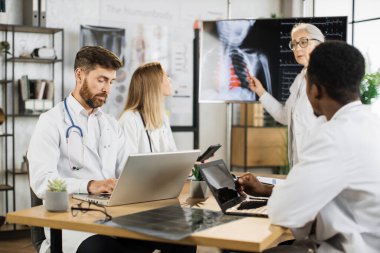 Beyaz önlüklü, beyaz önlüklü beyaz bir kadın uzman uluslararası doktorlarla brifing sırasında büyük monitörün yanında duruyor ve kalp atışları hızlanıyor. Tıp ve sağlık hizmetleri kavramı.