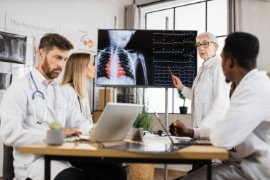 Beyaz önlüklü, beyaz önlüklü beyaz bir kadın uzman uluslararası doktorlarla brifing sırasında büyük monitörün yanında duruyor ve kalp atışları hızlanıyor. Tıp ve sağlık hizmetleri kavramı.