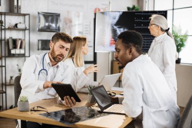 İki erkek doktor masada oturup tabletle dizüstü bilgisayar kullanırken kadın uzmanlar dijital monitörde röntgen taramasını inceliyor. Bir grup uluslararası tıp çalışanı toplantı odasında toplanıyor. .