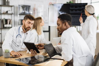 İki erkek doktor masada oturup tabletle dizüstü bilgisayar kullanırken kadın uzmanlar dijital monitörde röntgen taramasını inceliyor. Bir grup uluslararası tıp çalışanı toplantı odasında toplanıyor. .