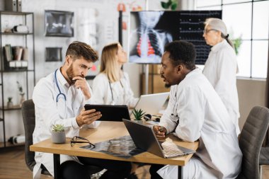 Laptop ve tabletle klinikteki masada otururken sohbet eden becerikli erkek tıp çalışanları. Laboratuvar önlüklü kadın doktorlar büyük dijital ekrandaki röntgen taramasını inceliyor..