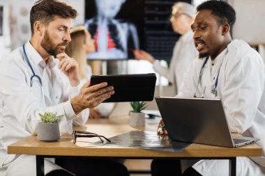 Kafkas ve Afrikalı Amerikalı erkek doktorlar arka planda bayan meslektaşlarıyla yaptıkları toplantıda tablet ve dizüstü bilgisayarla çalışıyorlar. Klinikteki uluslararası terapistlerin sabah brifingi.