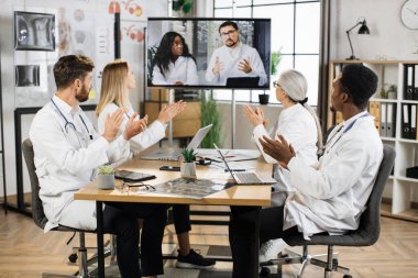 Farklı yaşlardan dört farklı bilim adamı toplantı odasındaki ortaklarla video konferansı sırasında el çırpıyor. Başarılı başarı, işbirliği ve tıp konsepti.