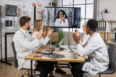 Heyecanlı kadın bilim adamının başarılı konuşmasından sonra online video konferansı sırasında beyaz laboratuvar önlüklü çok kültürlü doktorlar el çırpıyor. Ekrandaki Afrikalı Amerikalı kadına odaklan..
