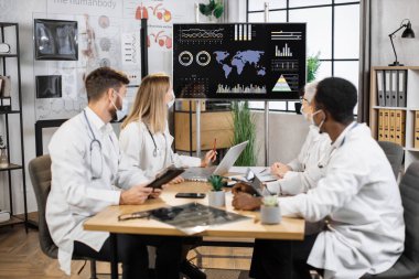 Hastane yönetim kurulu toplantısında tablet ve dizüstü bilgisayarlarla çalışan koruyucu maskeli çok ırklı çalışanlar. Doktorlar araştırmalarla monitöre bakarken koronavirüsün yayılmasını analiz ediyorlar..