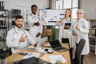 Uluslararası konferans sırasında kameraya aletlerle ve panoyla bakan gülümseyen doktorlar. Çok ırklı erkek ve kadın uzmanlar arka plandaki genetik formüller hakkında bilgi paylaşıyorlar..