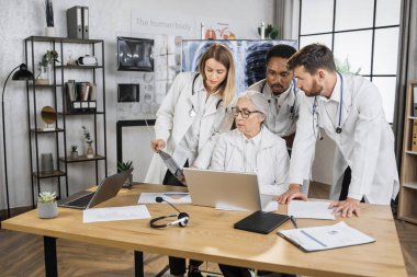 Üç stajyerden oluşan uluslararası ekip hastane konferans odasında kıdemli bayan doktorla acil bir toplantı yapıyor. Beyaz laboratuvar önlüklü işçiler hastaların tıbbi kayıtlarını incelemek için modern laptop kullanıyorlar..