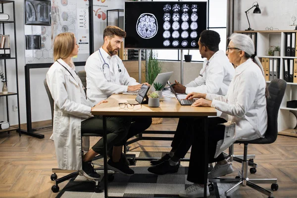 Team of international medical experts sitting at table and analyzing patients MRI scan on TV screen. Group of multiethnic male and female medical doctors using modern gadgets during work.