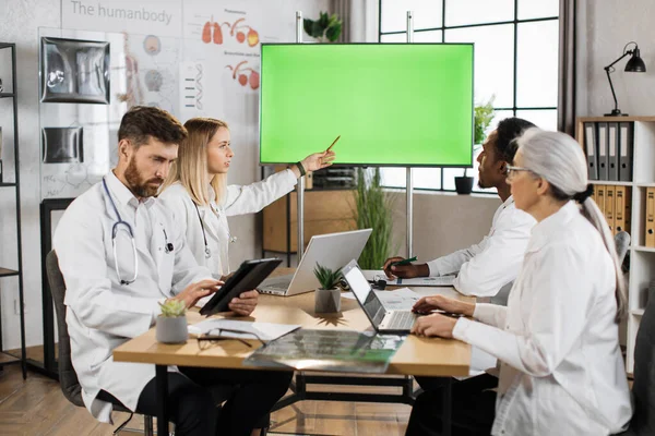 Team of multicultural medical specialists sitting at desk with modern gadgets and looking at big monitor with chroma key screen. Conference meeting of doctors at hospital office.
